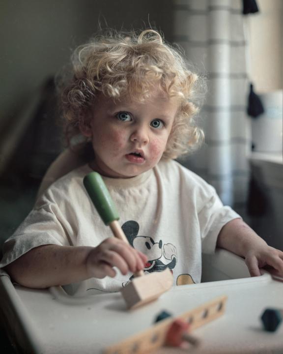 A child with curly blond hair and blue eyes sits in a high chair, holding a toy hammer.