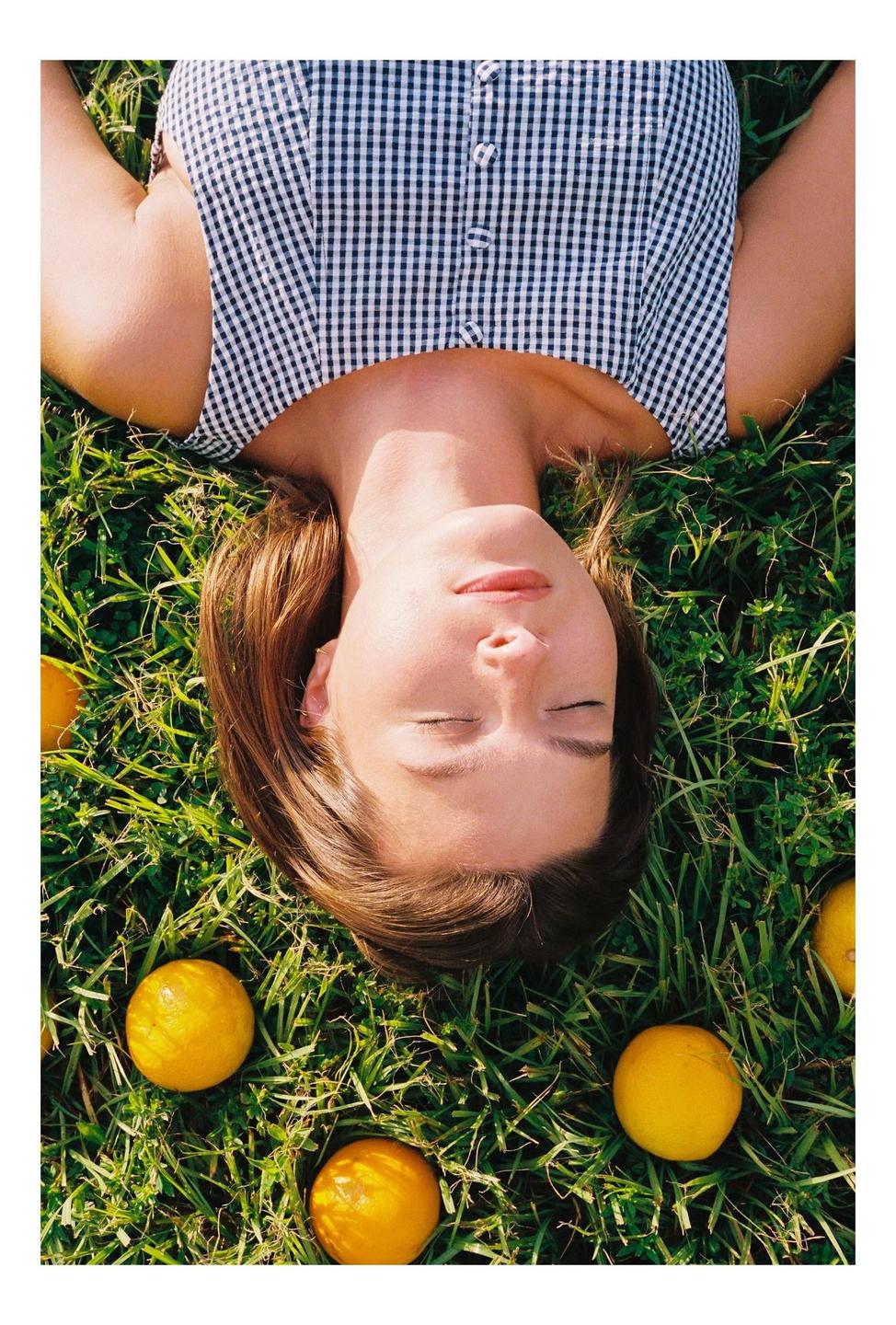 A woman with closed eyes lies in green grass surrounded by oranges.