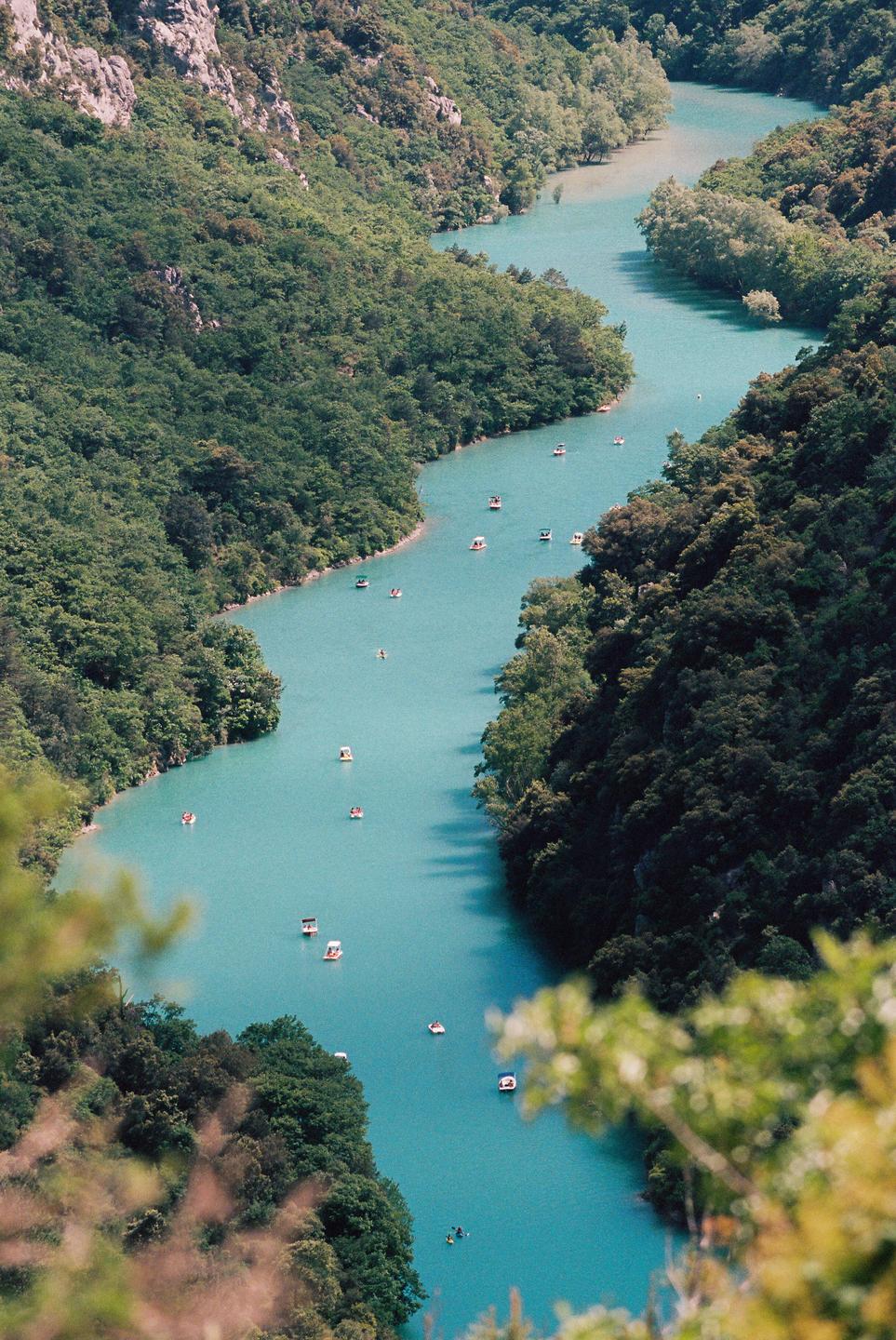 A winding turquoise river with many small boats is surrounded by lush green mountains.