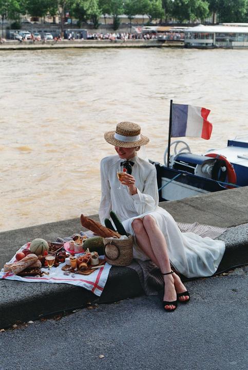 A woman in a white dress and straw hat sits by a river with a picnic spread, a French flag visible on a boat nearby.