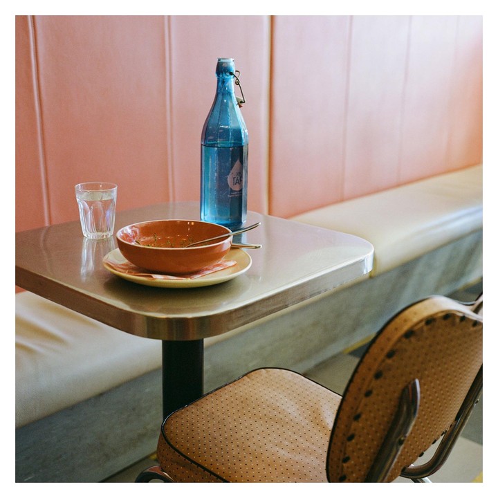 A blue bottle, a glass of water, and a bowl with a spoon are on a table next to a booth and a chair.