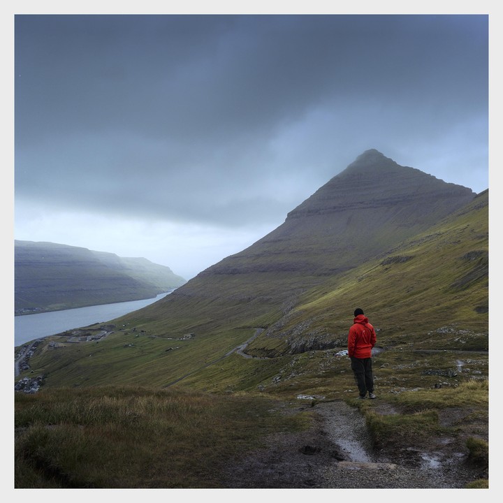 A person in a red jacket stands on a hill overlooking a fjord and a village nestled between mountains under a cloudy sky.