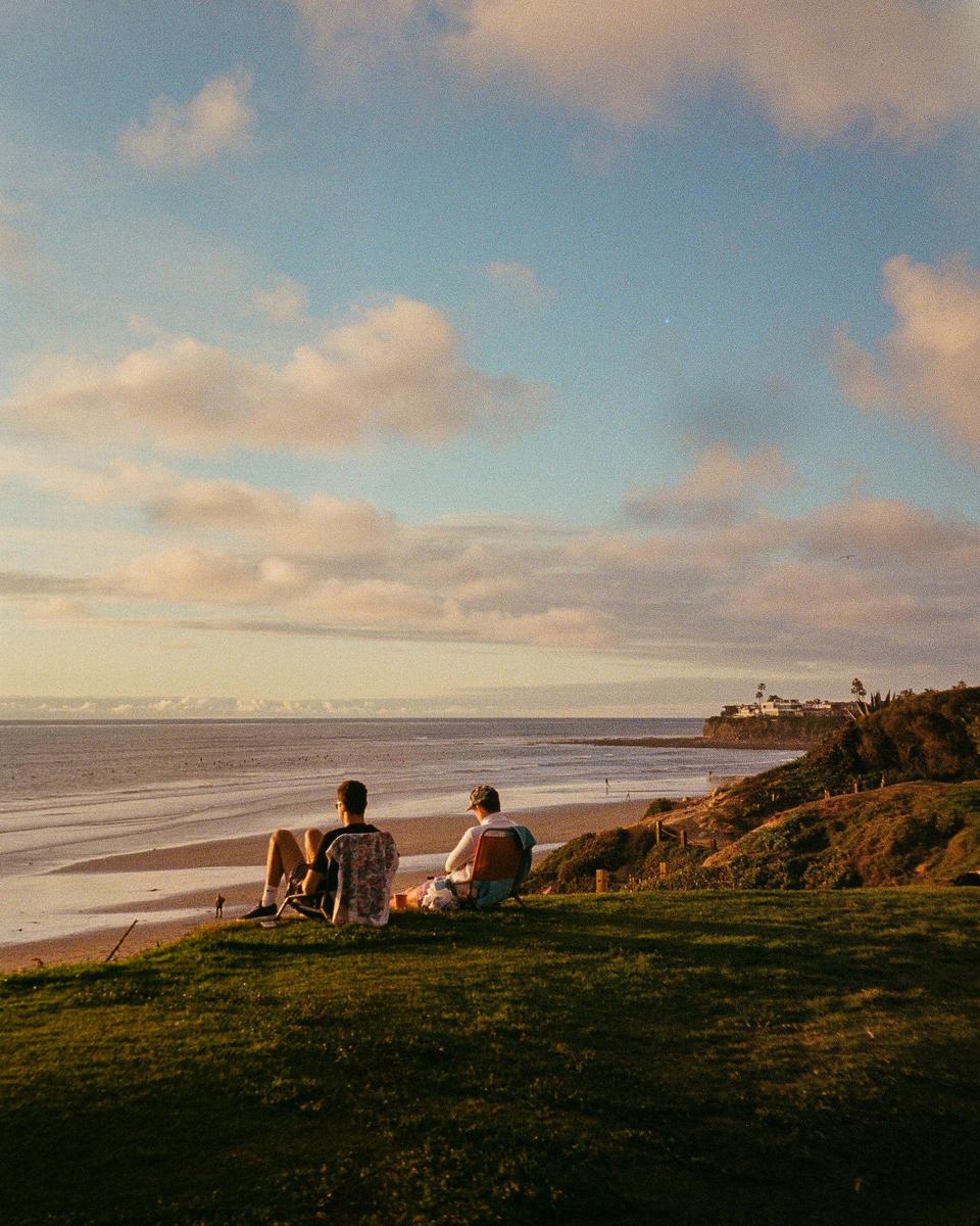 Two men sit on a grassy cliff overlooking a beach and ocean at sunset.