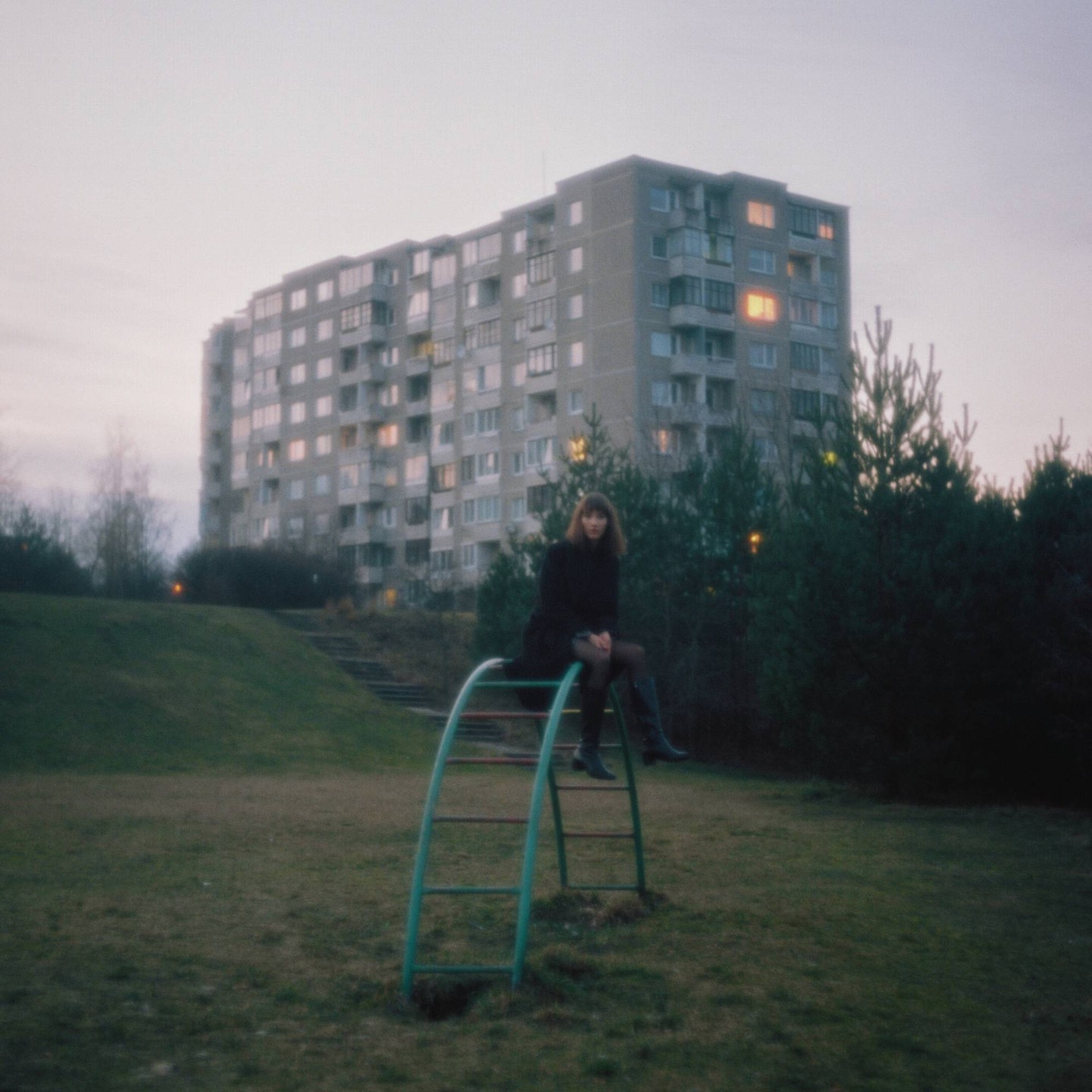 A woman sits on a metal climbing frame in a grassy area with a large apartment building in the background.
