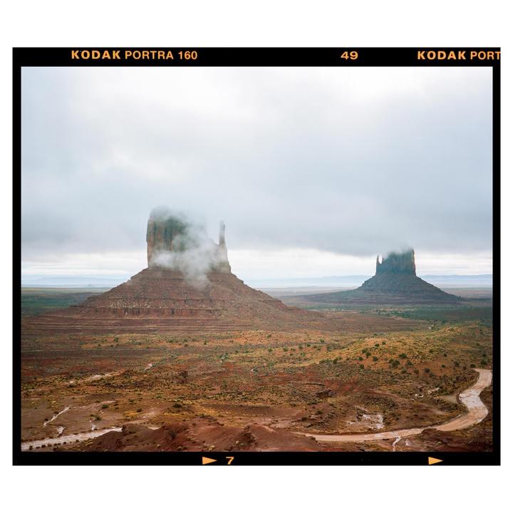 Two large rock formations rise from a desert landscape under a cloudy sky, with mist partially obscuring one of the formations.