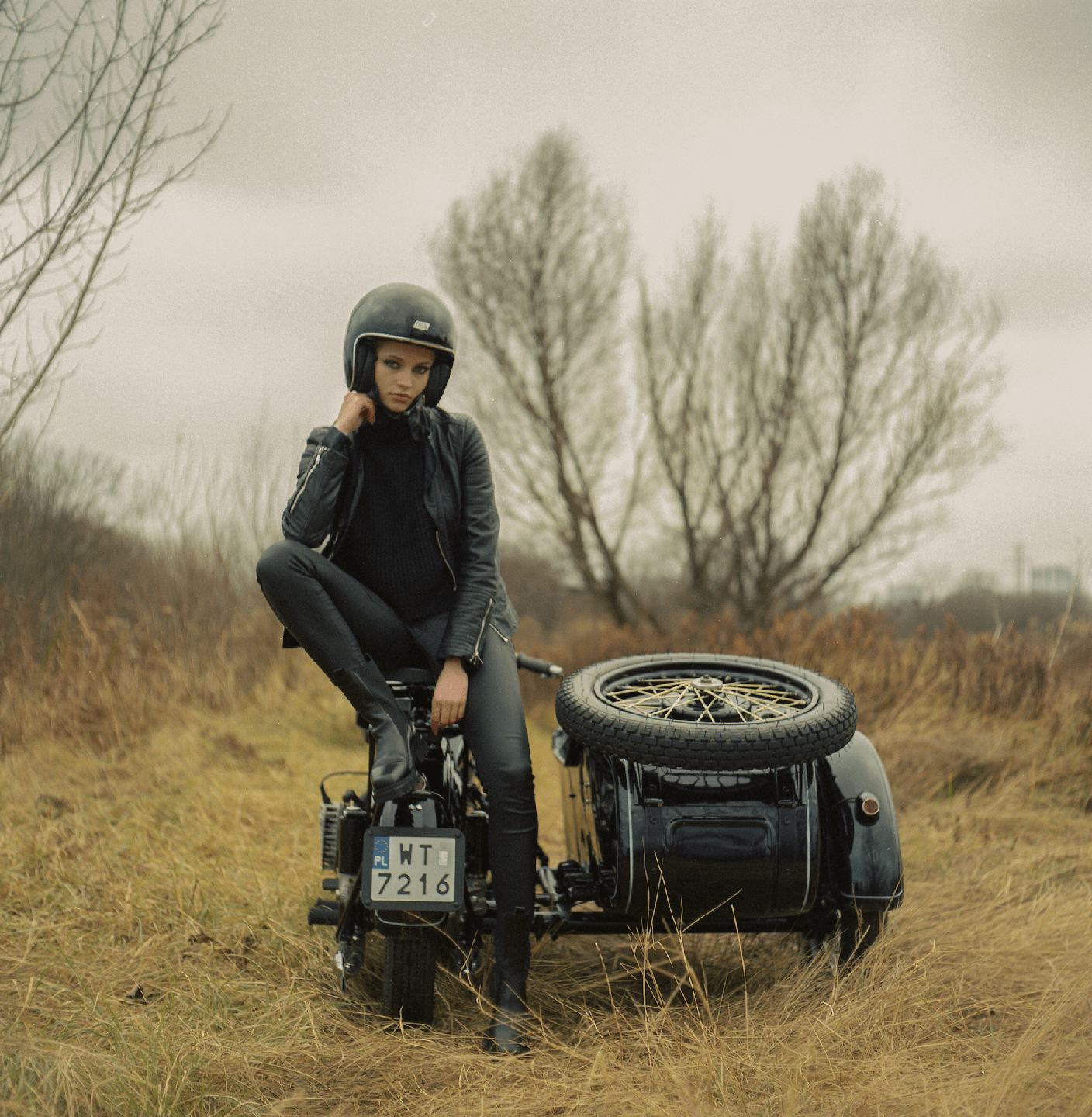 A woman in a helmet and leather jacket sits on a motorcycle with a sidecar in a field.