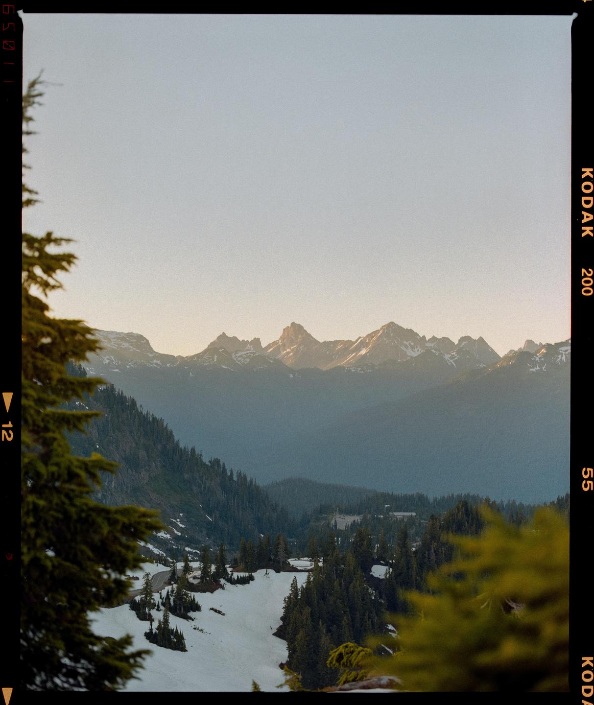 A mountain range with snow-covered slopes and evergreen trees is visible under a clear sky.