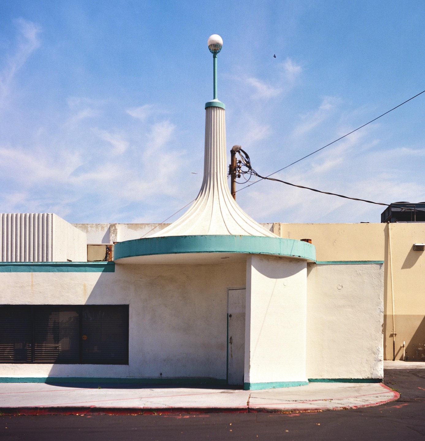 A white building with a teal trim and a tall, fluted spire with a spherical light on top stands under a blue sky.