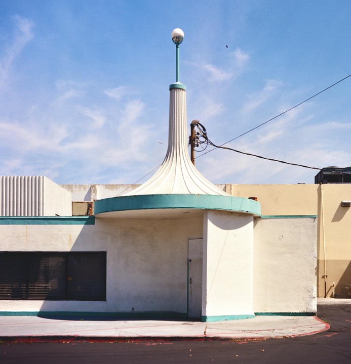 A white building with a teal trim and a tall, fluted spire with a spherical light on top stands under a blue sky.