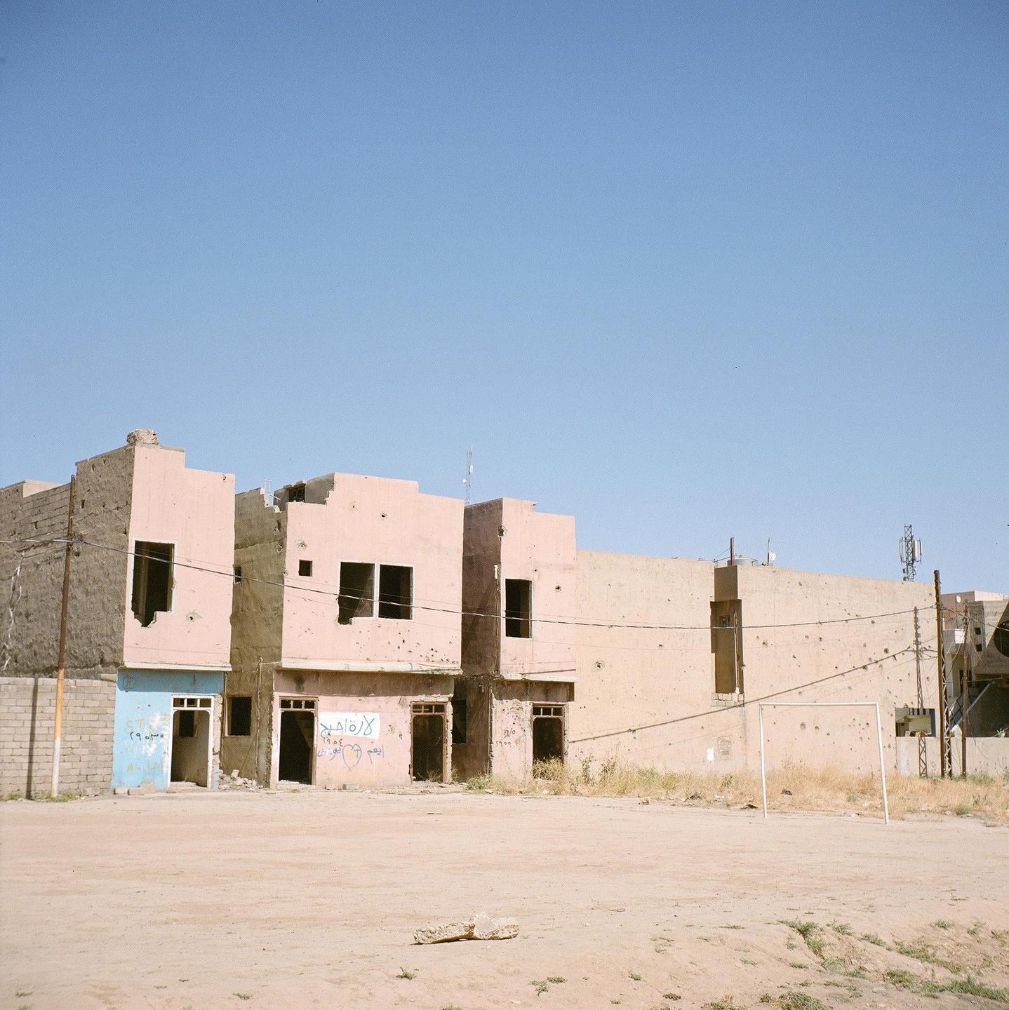 A soccer goal stands in a dirt field in front of bullet-ridden buildings under a clear sky.