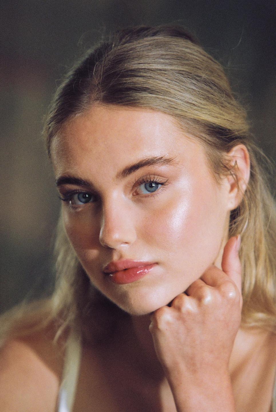 A woman with blonde hair and blue eyes looks directly at the viewer, resting her chin on her hand.