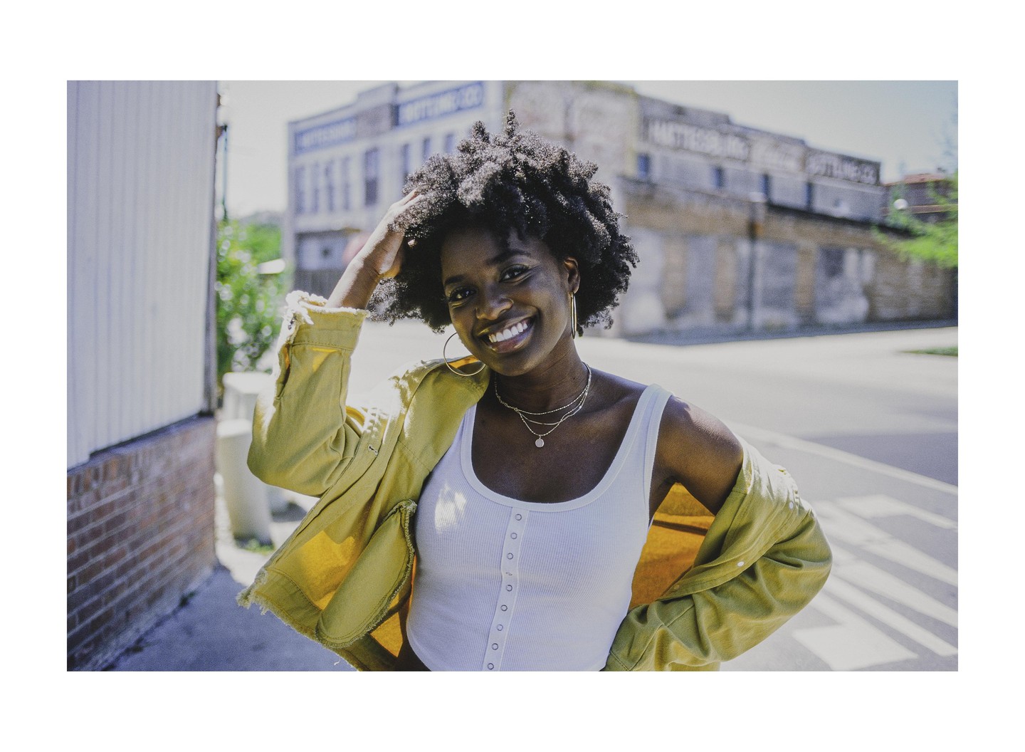 A woman with curly hair and a yellow jacket smiles at the camera on a street.