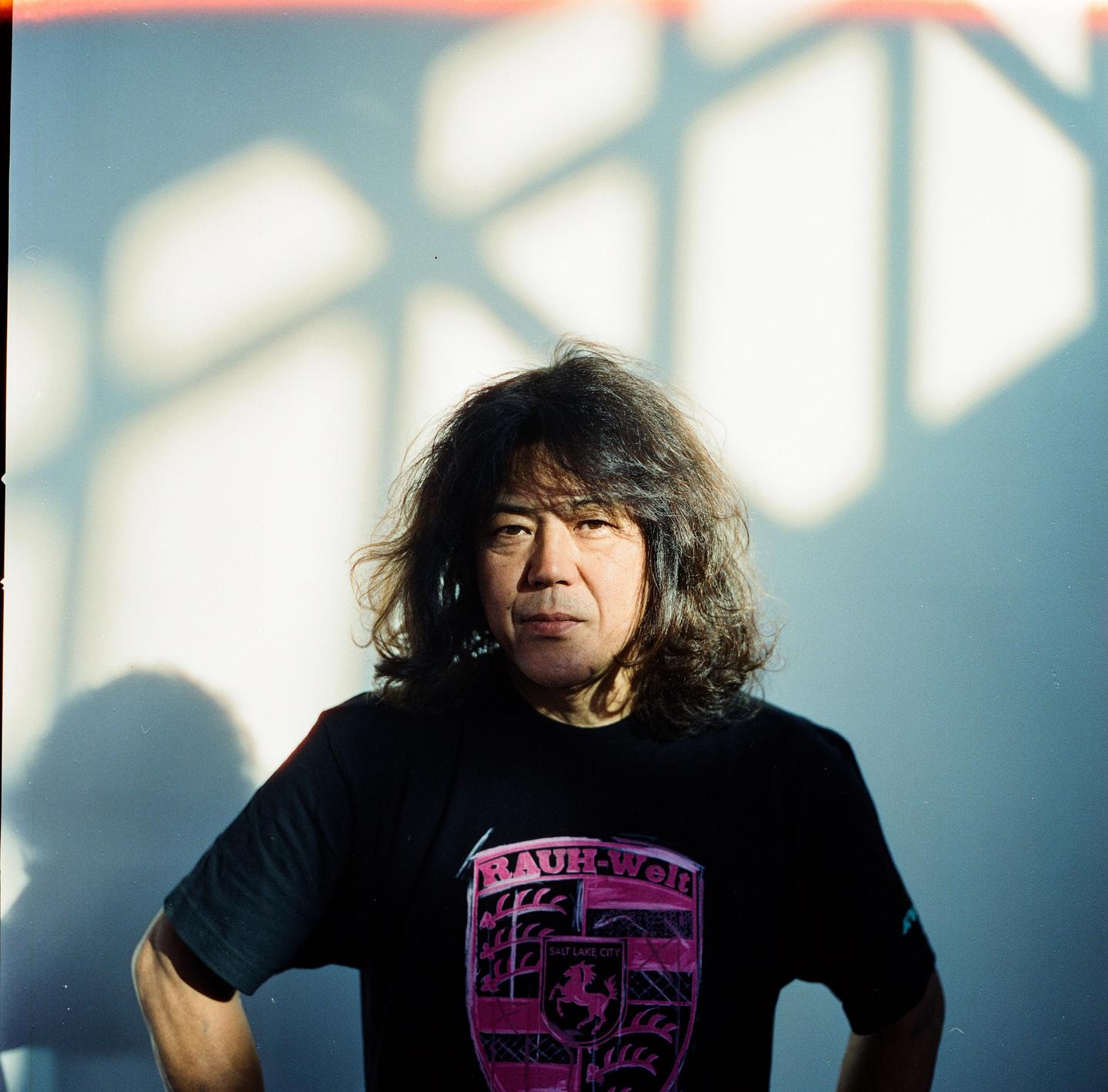 A man with long, dark hair and a black t-shirt stands in front of a wall with geometric shadows.
