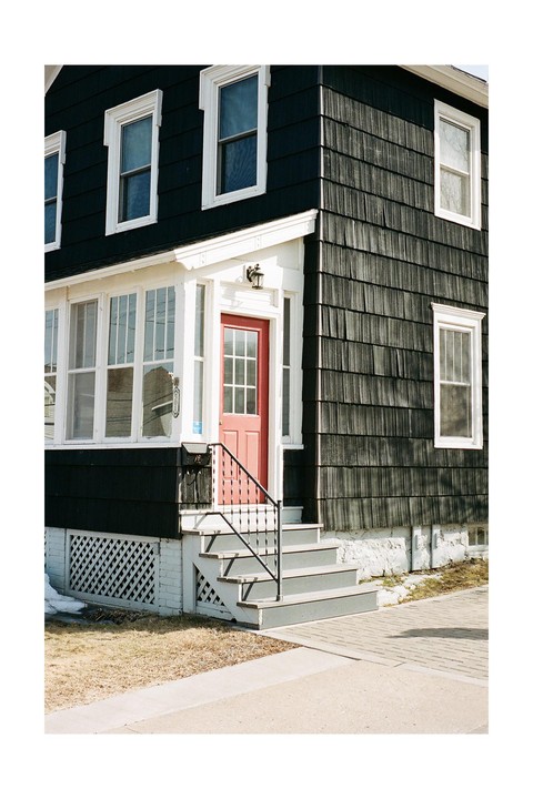 A black house with a red door and white trim stands next to a sidewalk with patches of dry grass and snow.