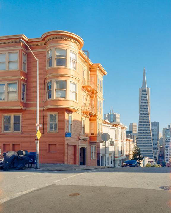 A peach-colored building with bay windows stands at a street corner, with a tall, pointed skyscraper visible in the distance.
