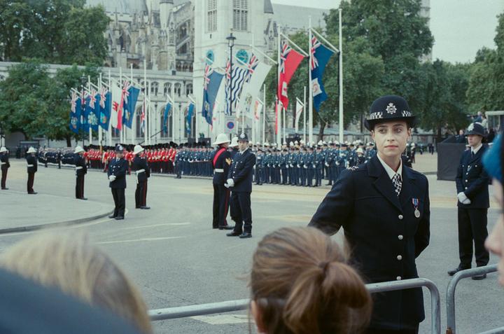 A woman in a police uniform stands in the foreground, with other uniformed officers and flags in the background.