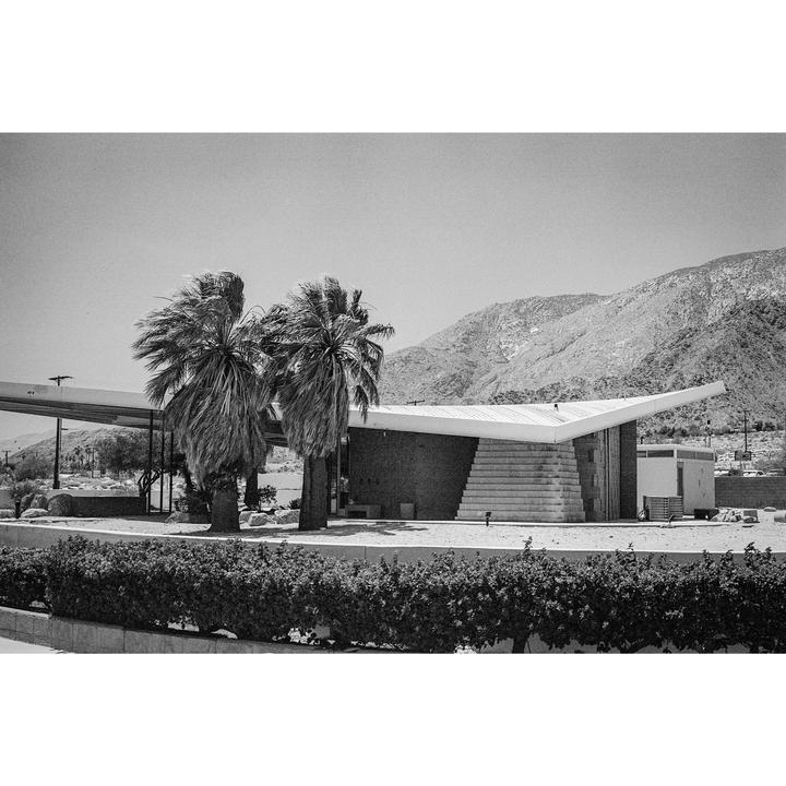 Two palm trees stand in front of a mid-century modern building with a mountain in the background.