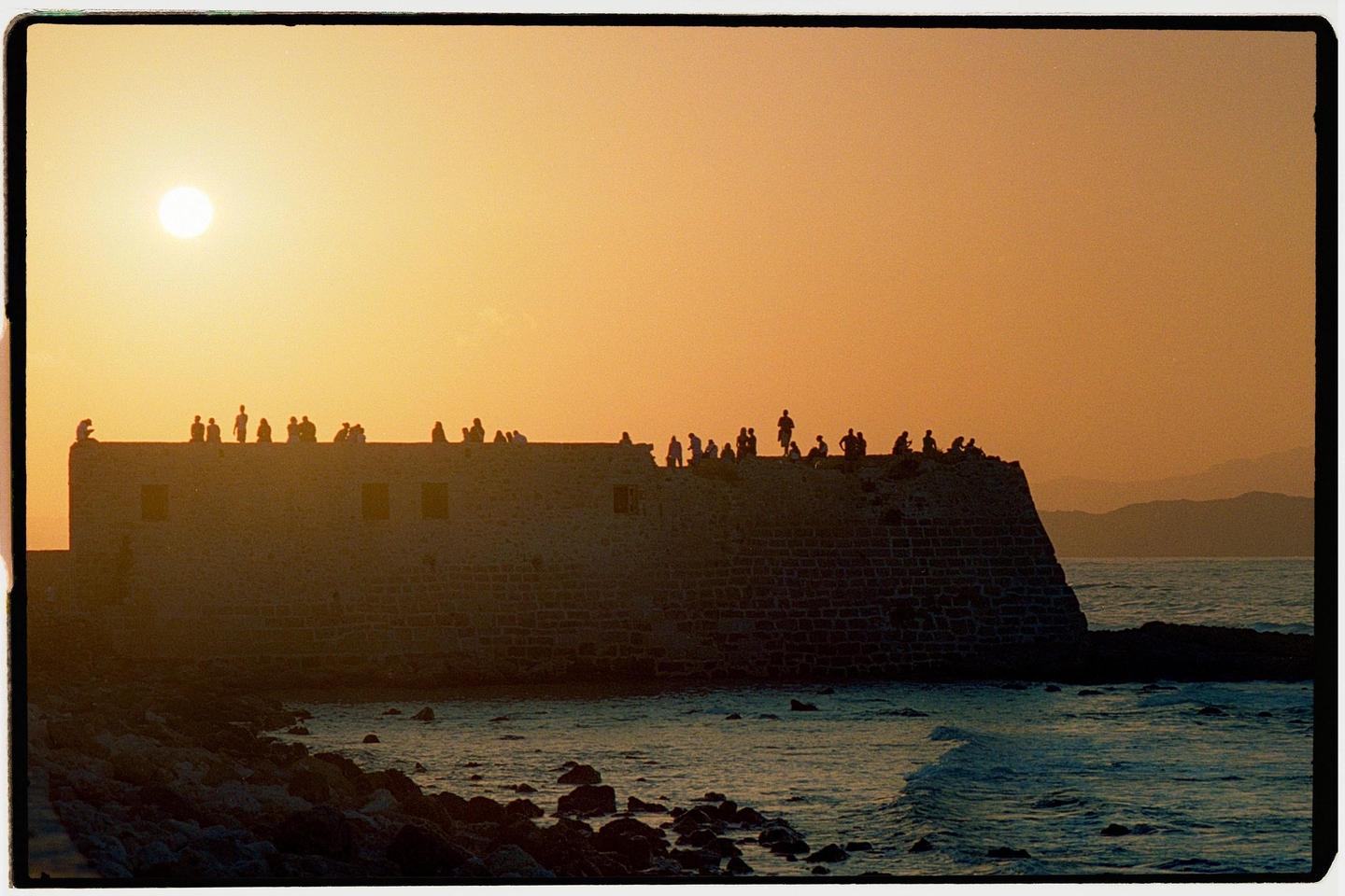 A group of people are silhouetted on a stone wall overlooking the sea at sunset.