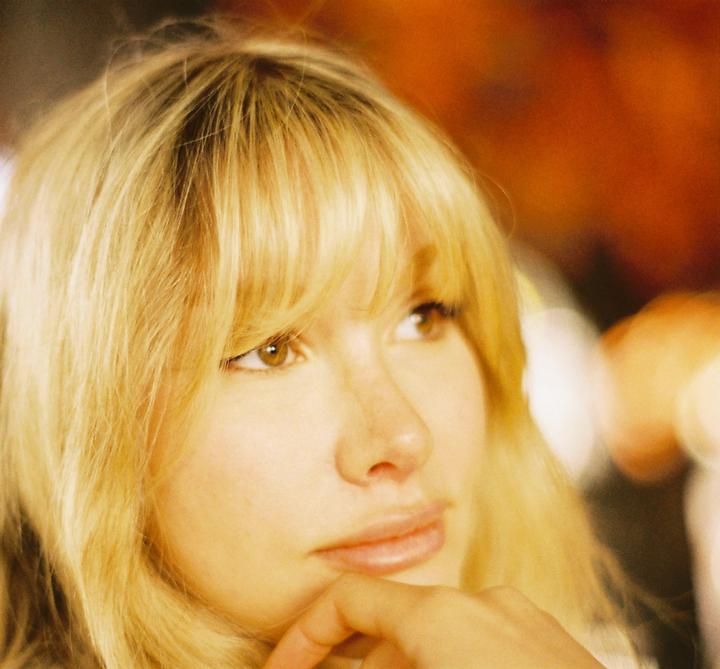 A woman with blonde hair and bangs looks to the side with her hand on her chin.