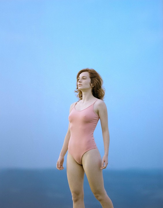 A woman in a pink swimsuit stands against a blue sky and ocean.