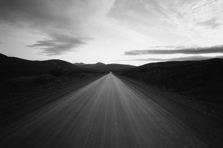 A long, straight dirt road stretches into the distance between rolling hills under a cloudy sky.