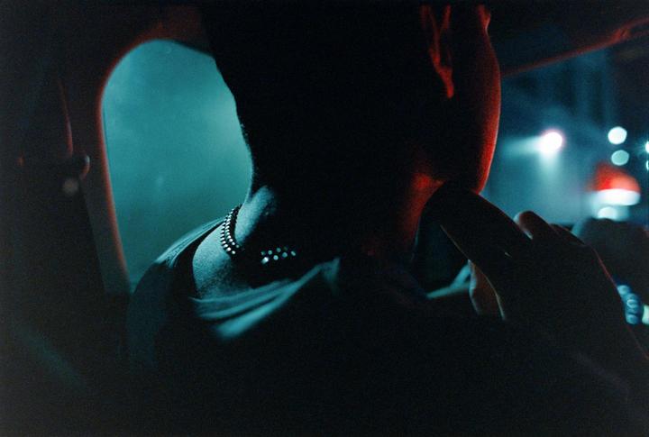 A person is visible from behind in a dark car interior, with a necklace visible and a hand near the face, while bright lights are visible outside the window.