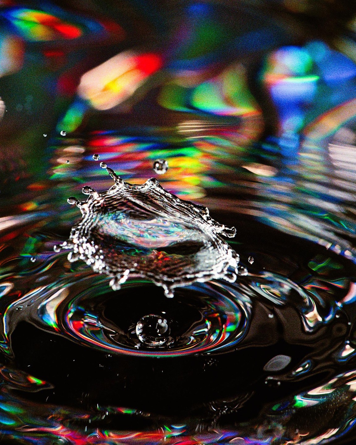 A water droplet creates a splash and ripples on a dark surface with colorful reflections.