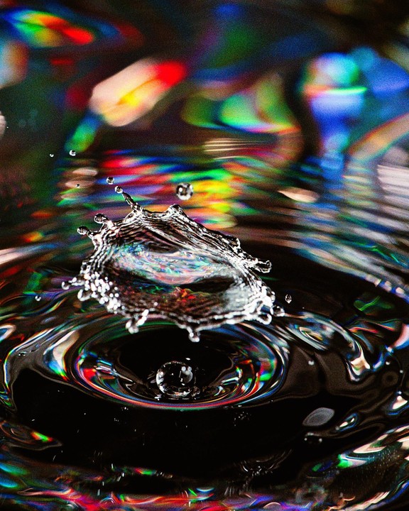 A water droplet creates a splash and ripples on a dark surface with colorful reflections.