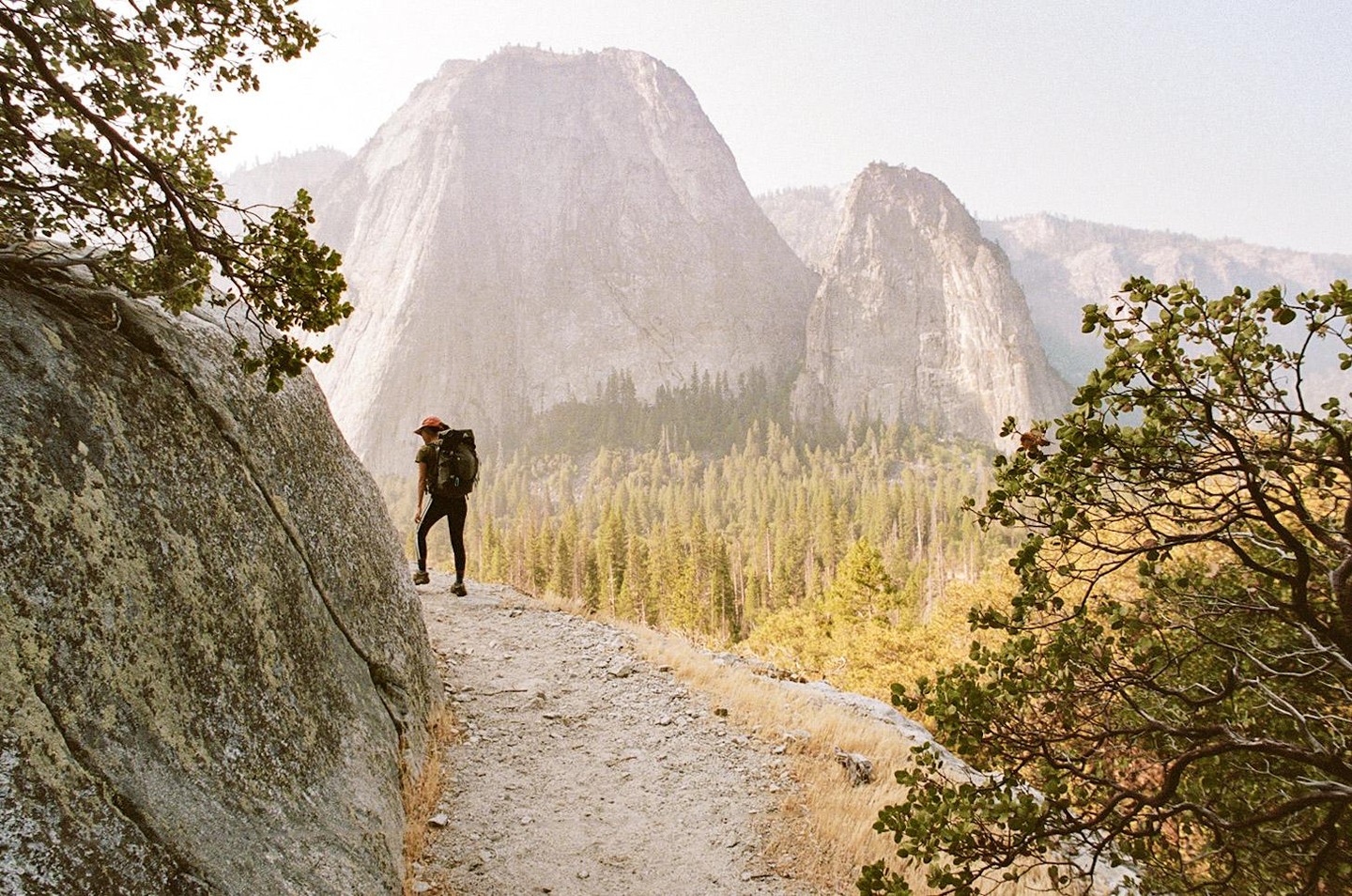 A person with a backpack walks on a dirt path next to a large rock formation, with a forest and mountains in the background.
