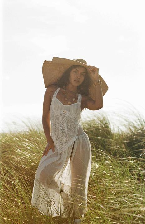 A woman wearing a large straw hat and a white dress stands in tall grass.