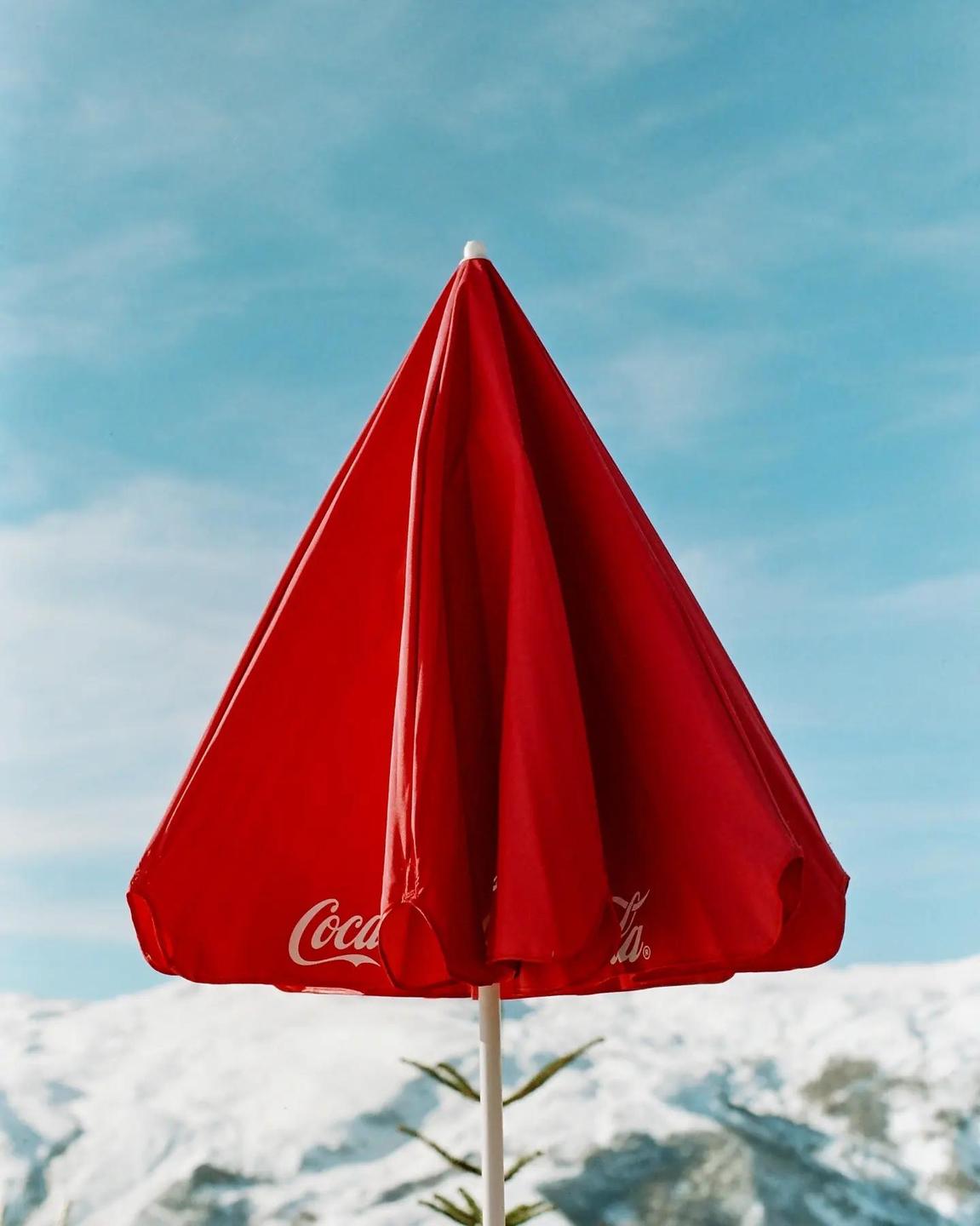 A red Coca-Cola umbrella stands against a clear blue sky with snow-covered mountains in the background.