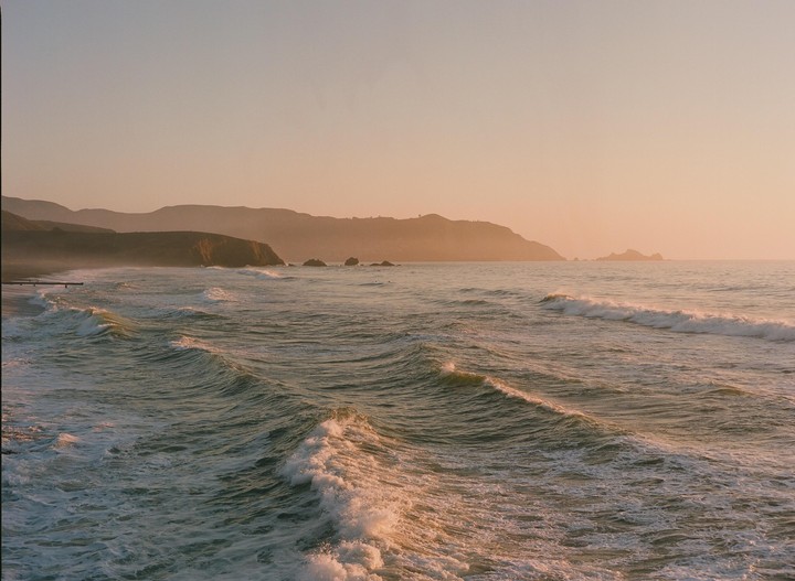 Waves roll towards a coastline with hills in the distance under a golden hour sky.