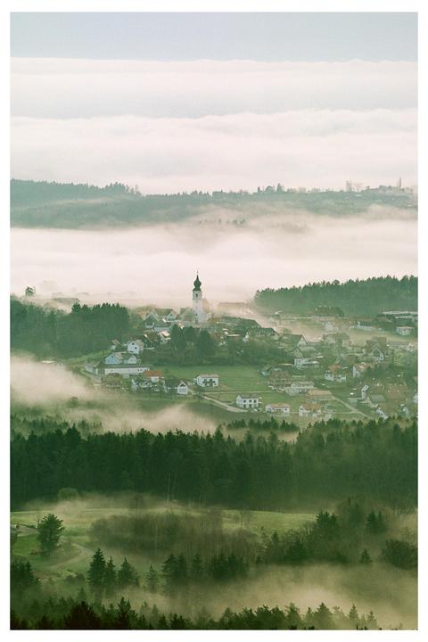 A village with a church steeple is nestled among rolling hills and forests, partially obscured by a layer of fog.