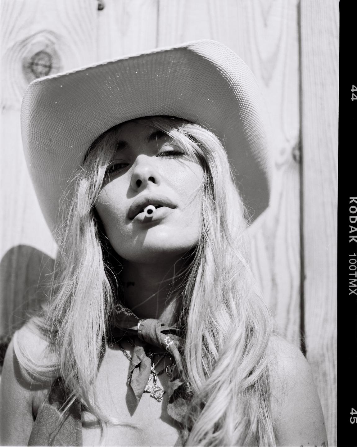 A woman wearing a cowboy hat and bandana holds a cigarette filter in her mouth.
