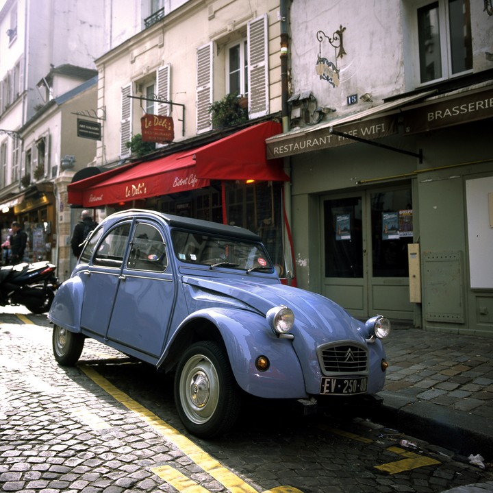 A light blue car is parked on a cobblestone street in front of a building with a red awning.