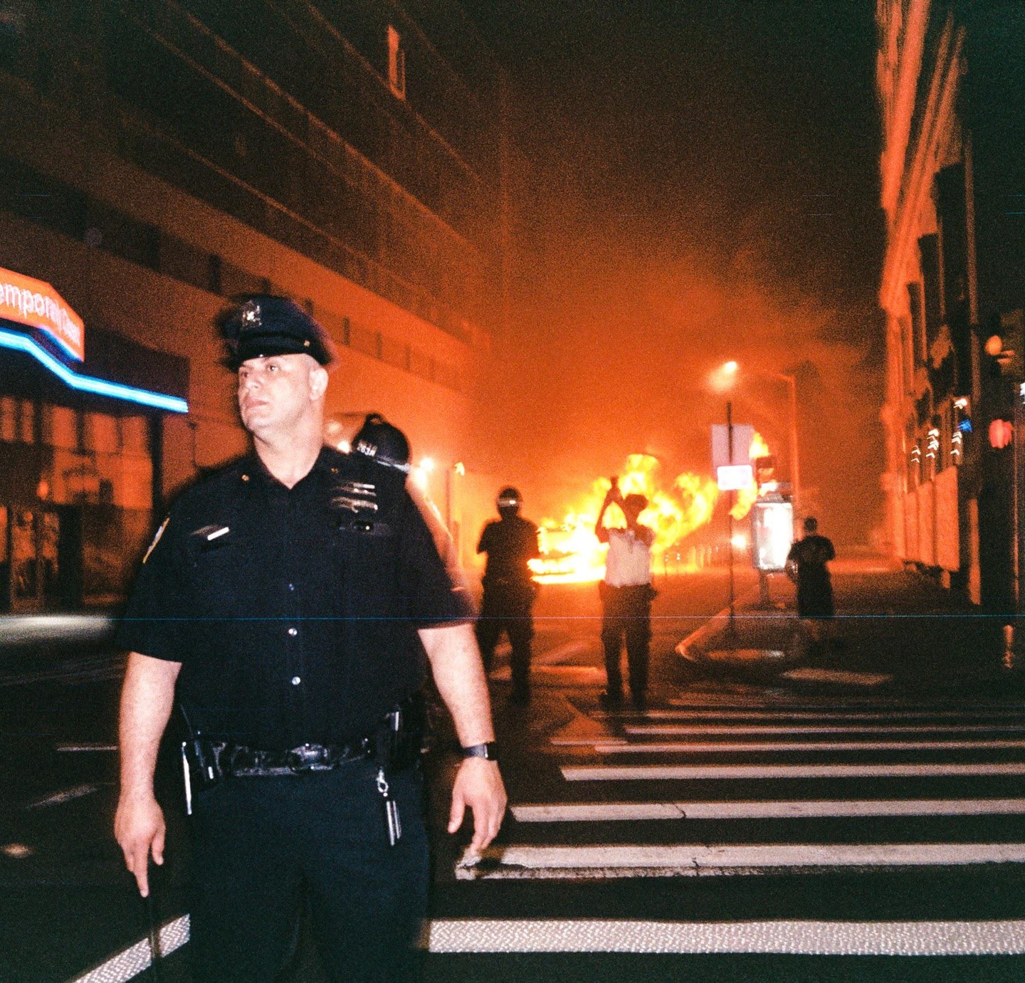 A police officer stands in the street at night with a large fire burning in the background.