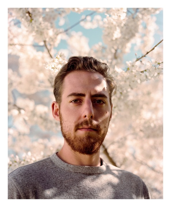 A man with a beard looks directly at the viewer, framed by white blossoms against a blue sky.