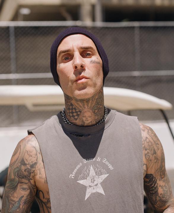 A man with numerous tattoos on his face, neck, and arms wears a dark beanie and a gray sleeveless shirt.
