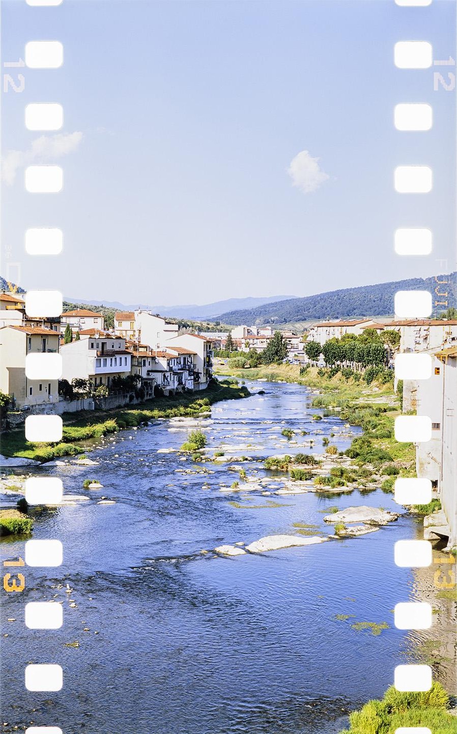 A river with rocks and vegetation flows through a town with white buildings and red roofs, surrounded by hills under a clear sky.