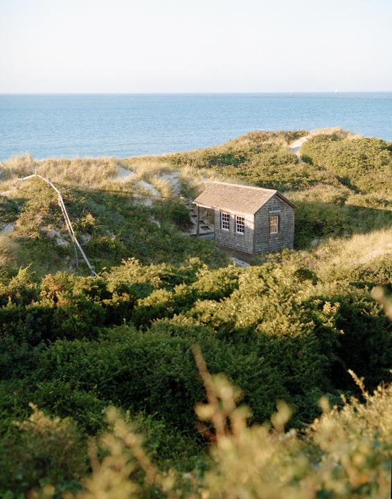 A small wooden house with a porch sits on a grassy dune overlooking the ocean.