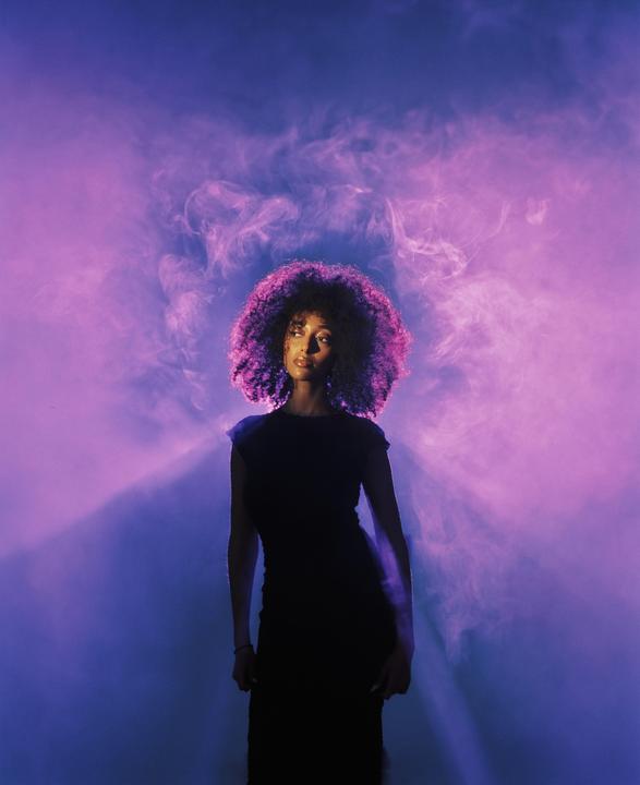 A woman with curly hair stands in a hazy, purple-lit environment.