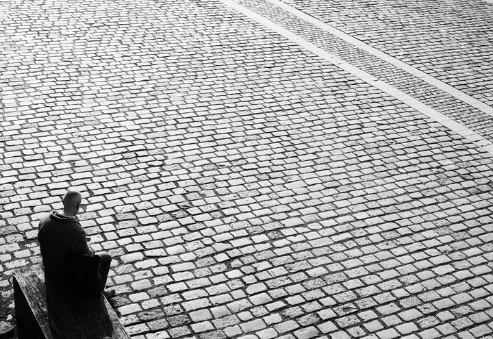 A man sits on a wooden bench looking out over a large expanse of cobblestone.