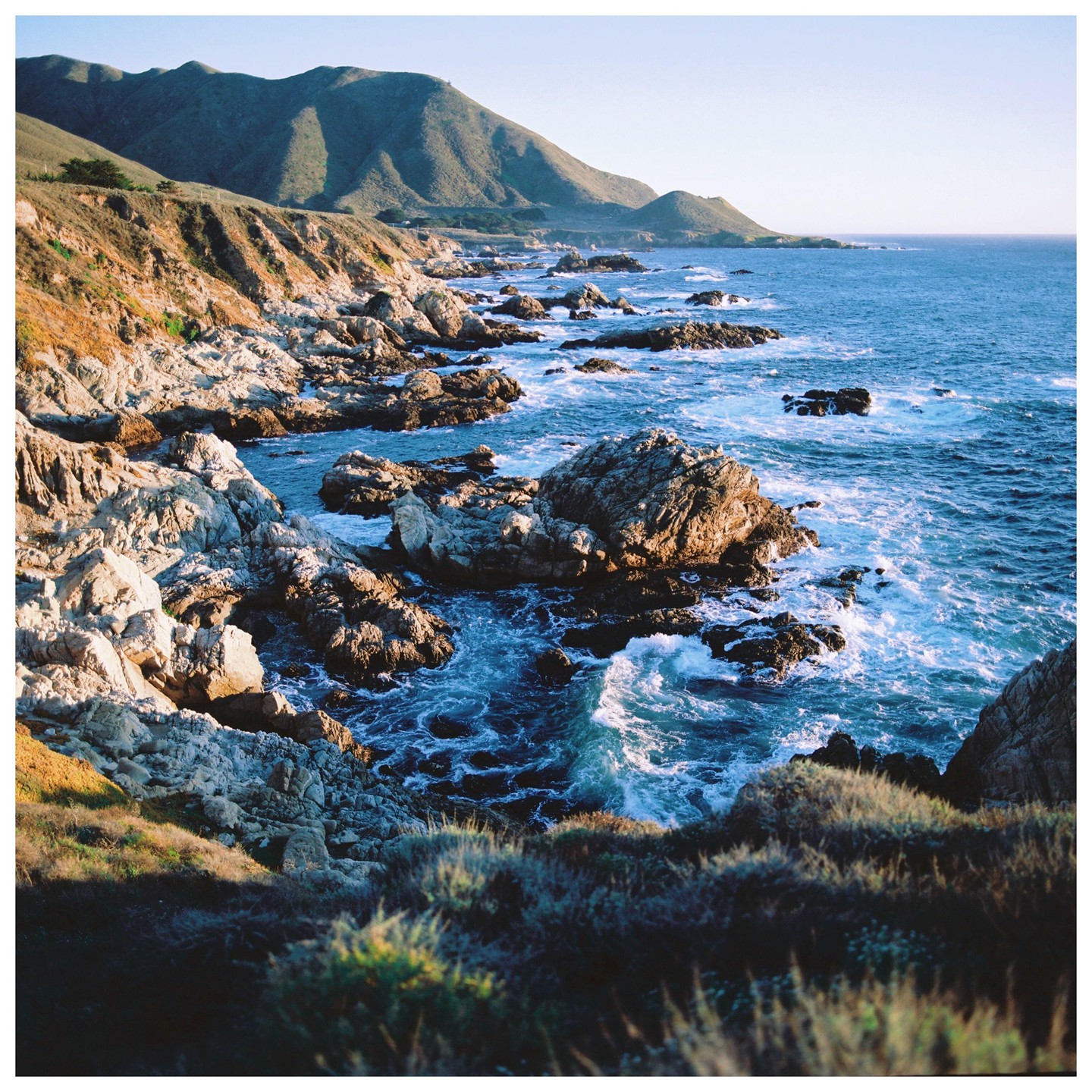 Waves crash against a rocky coastline with mountains in the background.