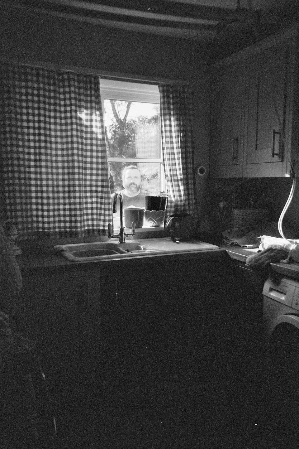 A man with a beard looks through a kitchen window with checkered curtains.