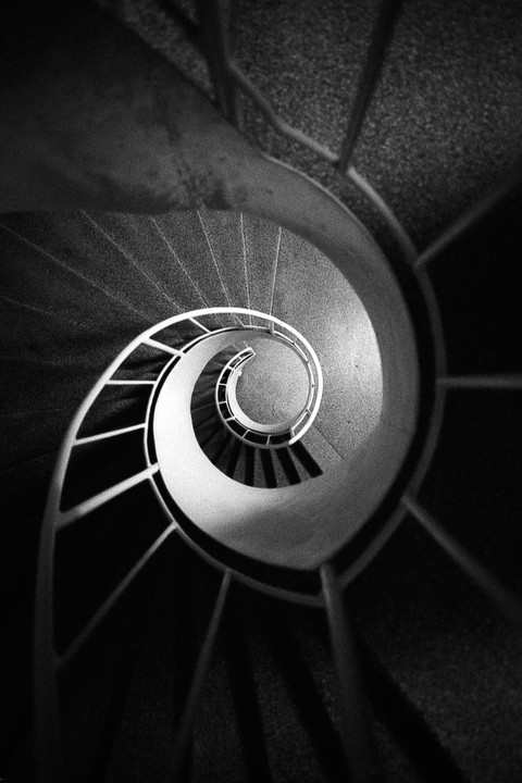 A spiral staircase with a white railing descends into darkness.