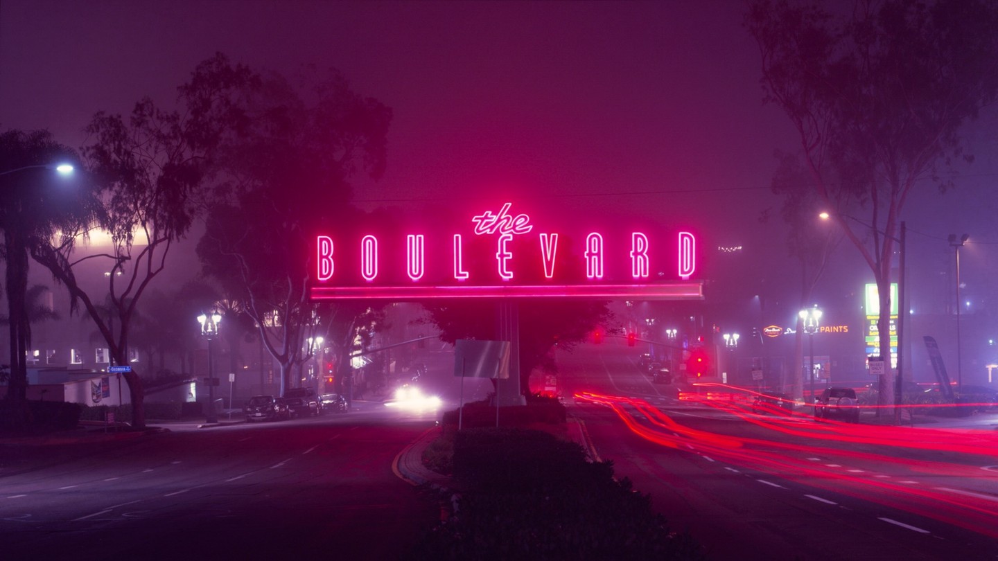 A neon sign for "the Boulevard" glows pink above a street at night with light trails from cars.