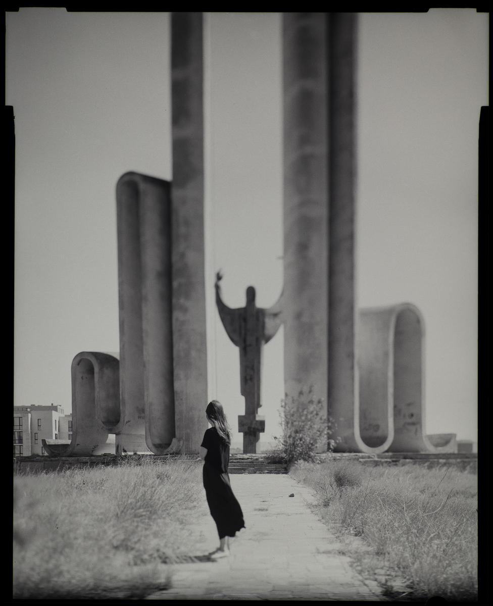 A woman stands barefoot on a path looking at a large concrete sculpture with other concrete structures in the background.