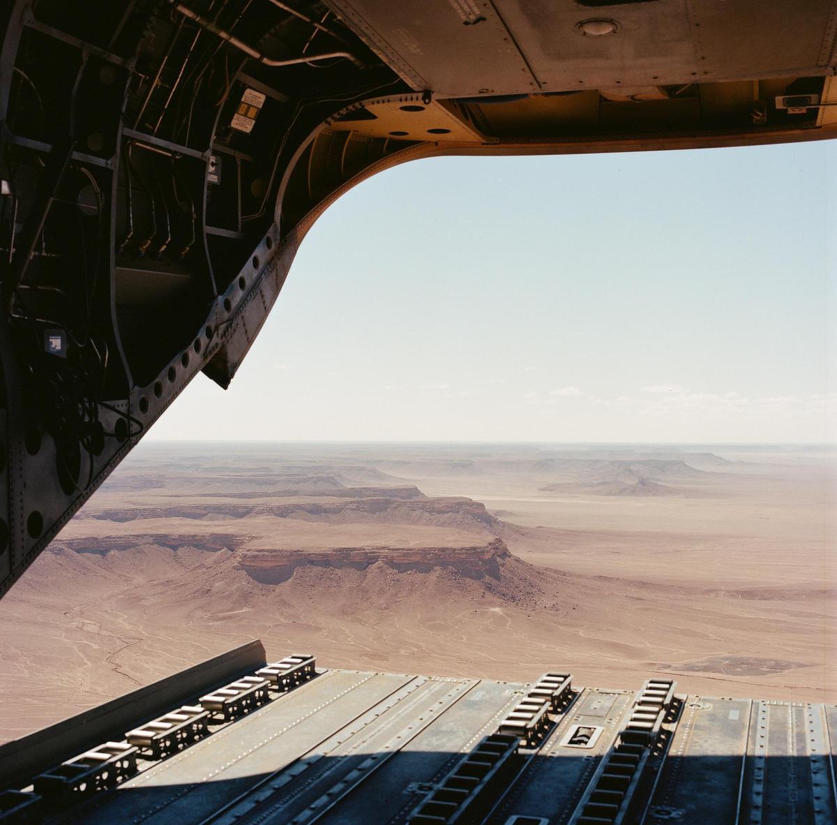 A view from the back of an aircraft shows a vast desert landscape with mesas and canyons under a clear sky.