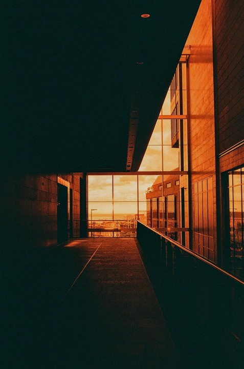 A dark interior hallway with a ramp leads to a large window overlooking a city and the ocean at sunset.