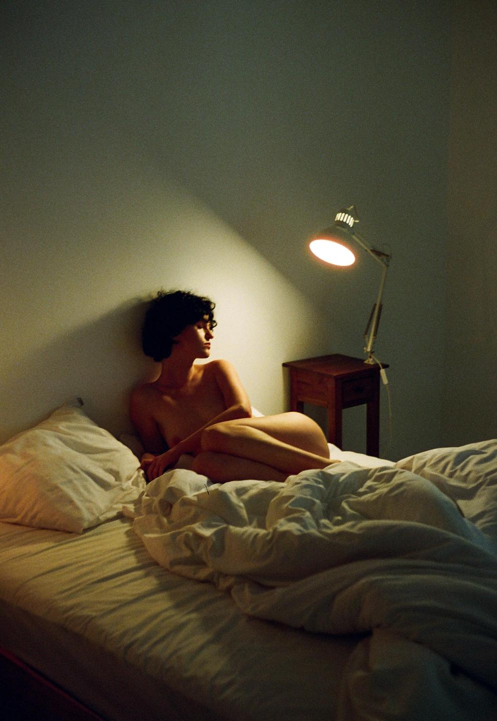A woman with short, curly hair is lying on a bed, illuminated by a lamp on a bedside table.